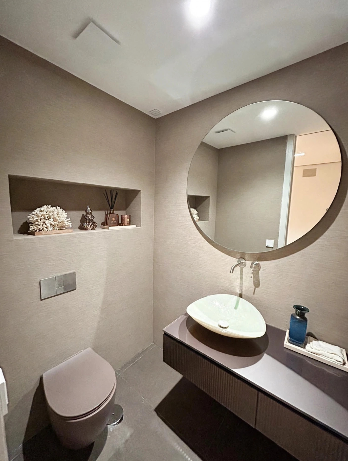 Baño de cortesía en tonos piedra con lavabo suspendido y espejo circular